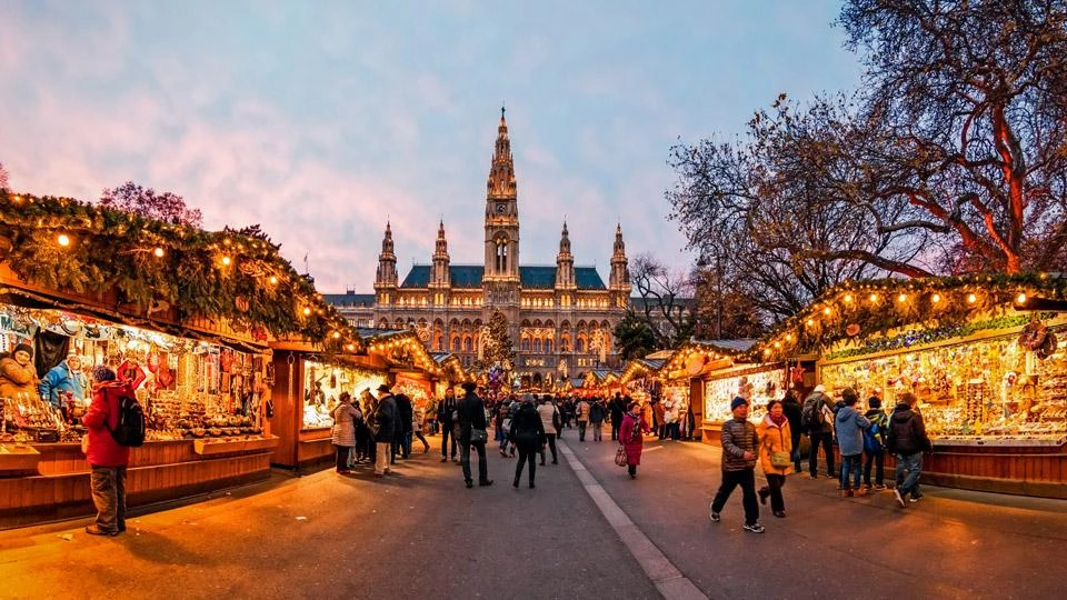Wooden Christmas market stalls glowing with lights in front of a Gothic town hall at dusk