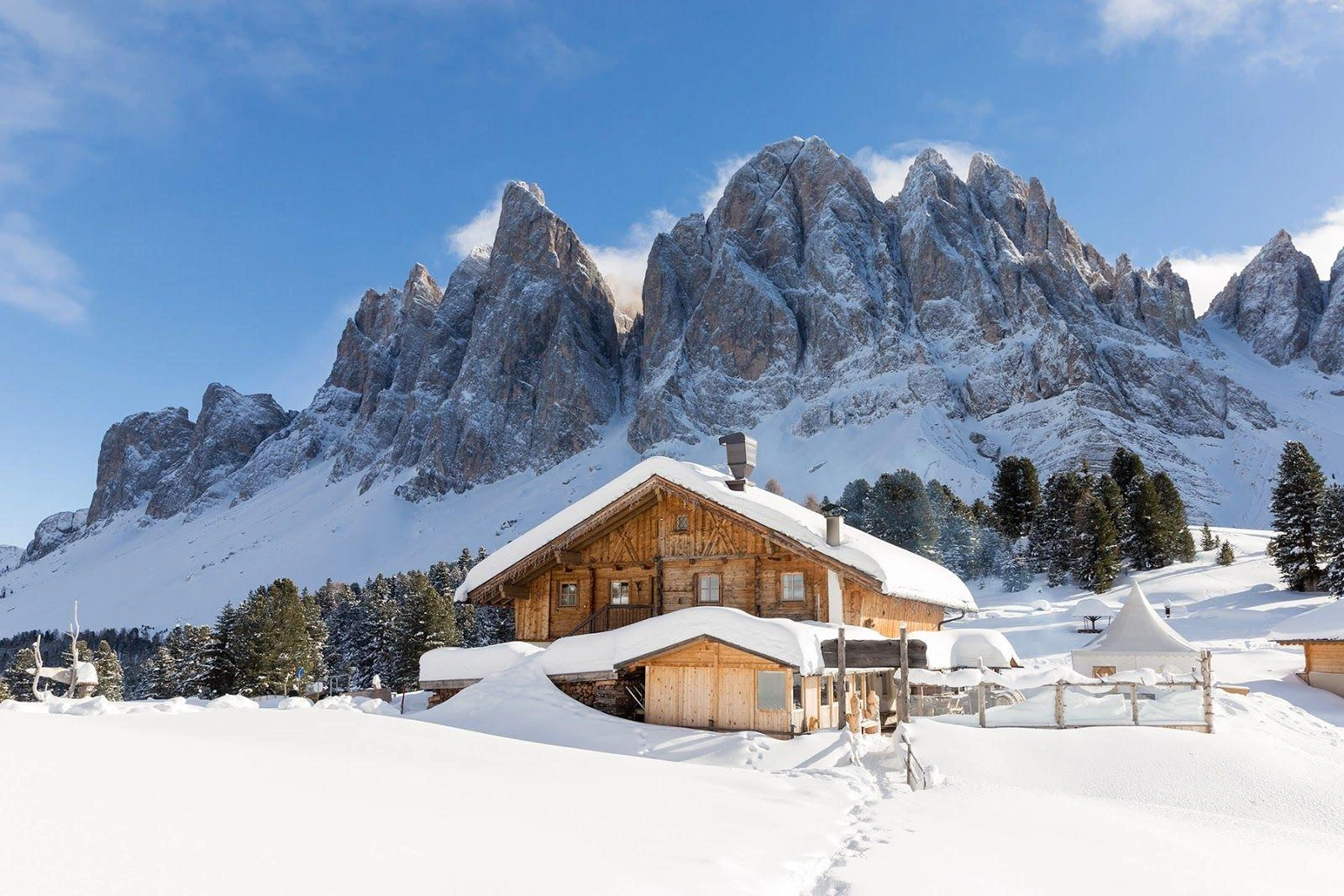 A snow-covered timber alpine hut below jagged Dolomite peaks on a clear winter day